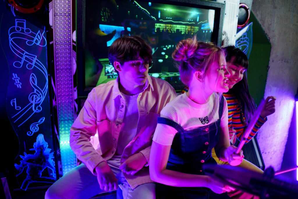 Three friends having fun at an arcade, playing music games with vibrant neon lights.